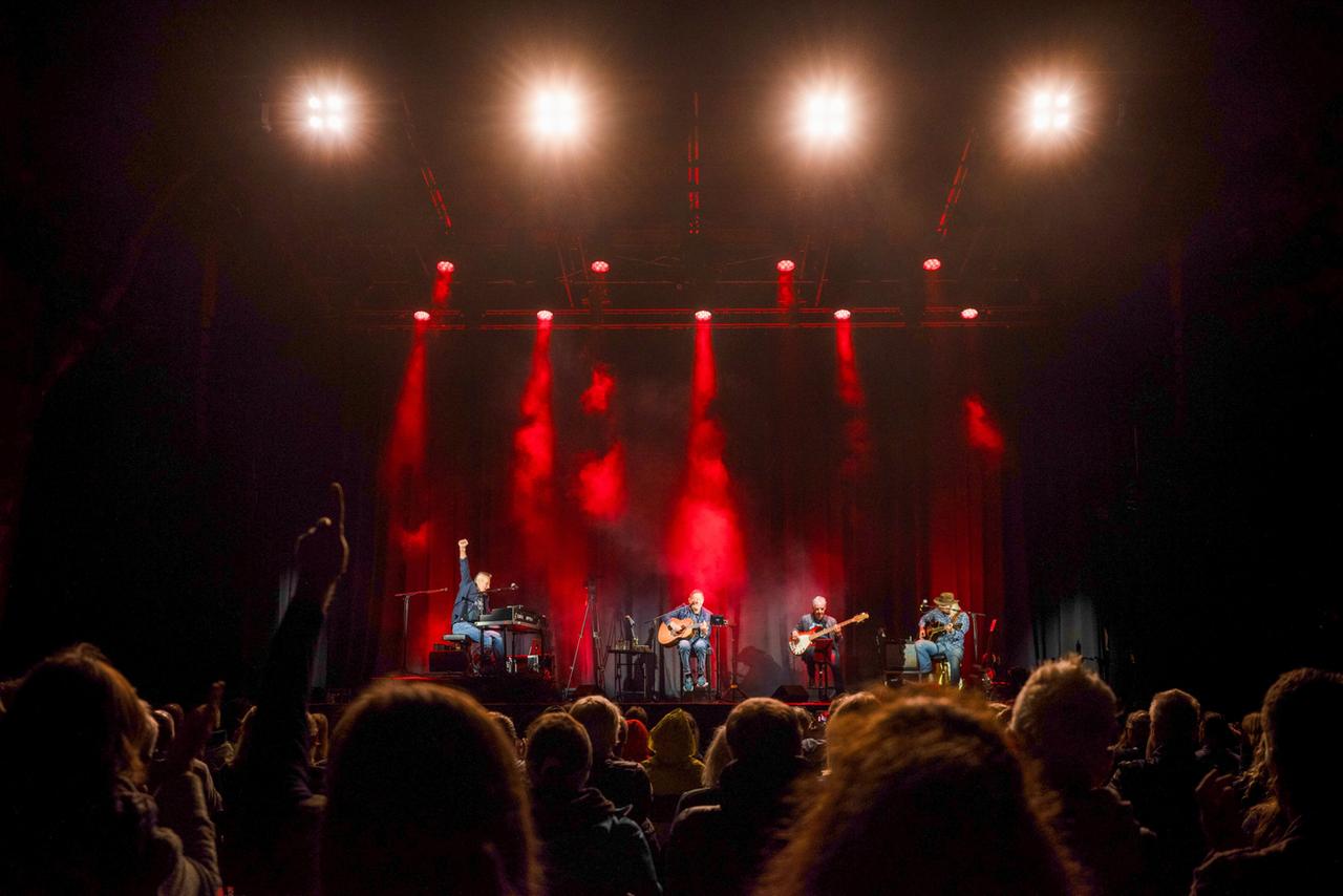 Das Bild zeigt eine Live-Performance auf einer Bühne im Theater im Park. Im Vordergrund sind mehrere Musiker zu sehen, die in konzentrierter Haltung Instrumente spielen. Ein Sänger, der am Keyboard sitzt, hebt eine Hand in die Luft, während er mit der anderen spielt. Zu seiner linken sitzt ein weiterer Musiker mit einer akustischen Gitarre, während zwei andere Musiker im Hintergrund einen Bass und eine elektrische Gitarre spielen. Die Bühne ist mit rotem und weißem Licht beleuchtet, das von oben herabstrahlt, was eine festliche Atmosphäre schafft. Im Vordergrund sind die Köpfe des Publikums zu sehen, das auf den Auftritt reagiert. Das gesamte Setting vermittelt ein Gefühl von Begeisterung und Gemeinschaft unter den Zuschauern.