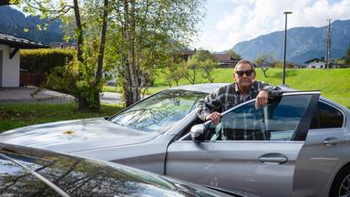 Das Bild zeigt einen älteren Mann, der an einem silbernen Auto steht. Er trägt eine kariierte Jacke und eine Sonnenbrille. Der Mann hat eine entspannte Haltung und lehnt an der offenen Tür des Fahrzeugs.   Im Hintergrund sind grüne Wiesen und Bäume zu sehen, die von der herbstlichen Färbung geprägt sind. Einige Häuser sind leicht unscharf im Hintergrund zu erkennen, und eine Berglandschaft ist ebenfalls sichtbar. Der Himmel ist blau mit einigen Wolken. Der gesamte Eindruck vermittelt eine ruhige ländliche Umgebung.