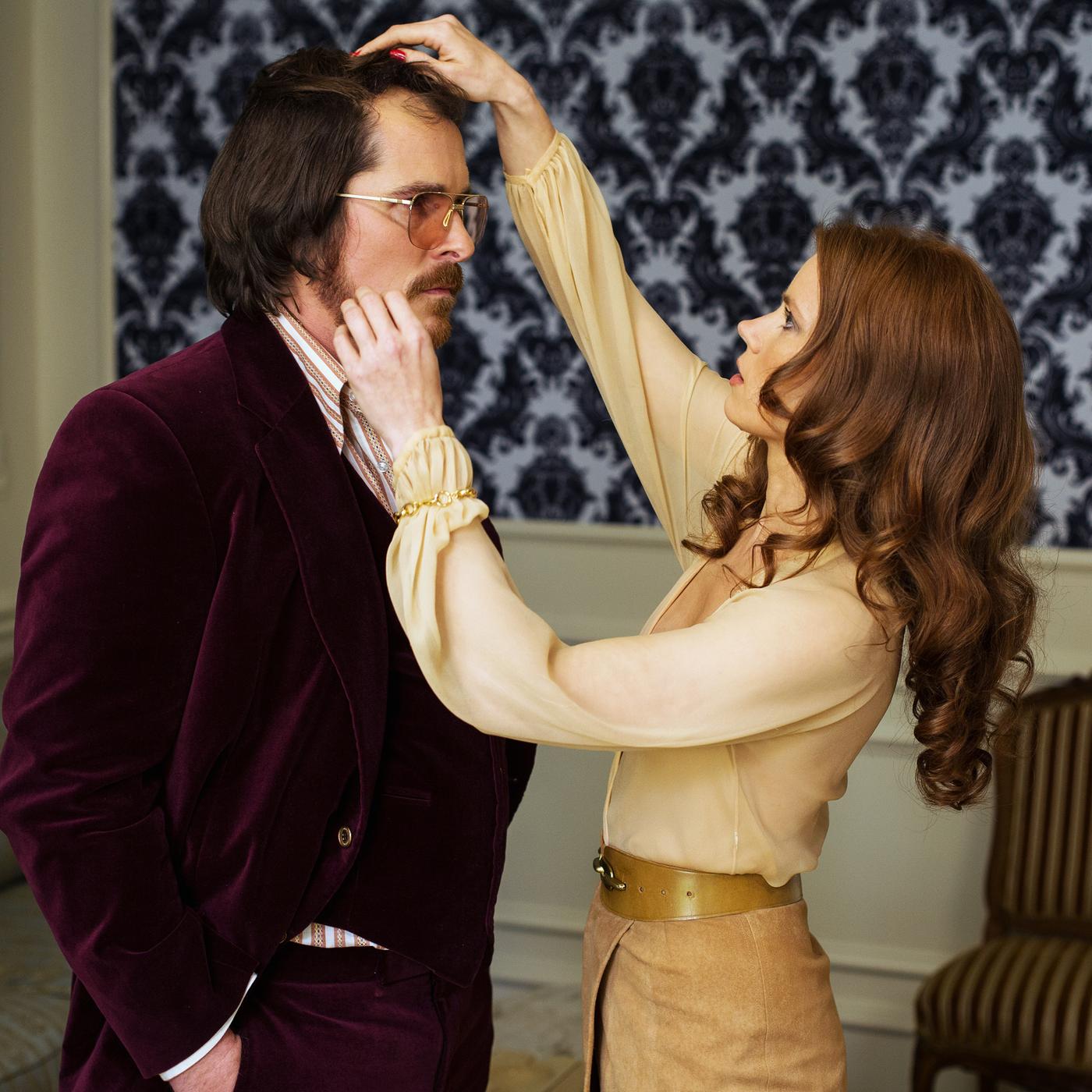 Szene aus American Hustle: Eine Frau in beigefarbener Bluse richtet einem Mann im bordeauxfarbenen Anzug das Toupé, während sie sich konzentriert auf ihn fokussiert.