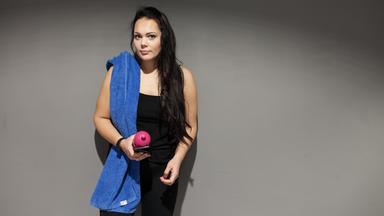 Eine junge Frau in Sportkleidung mit einem blauen Handtuch über der Schulter & einer Trinkflasche in der Hand.