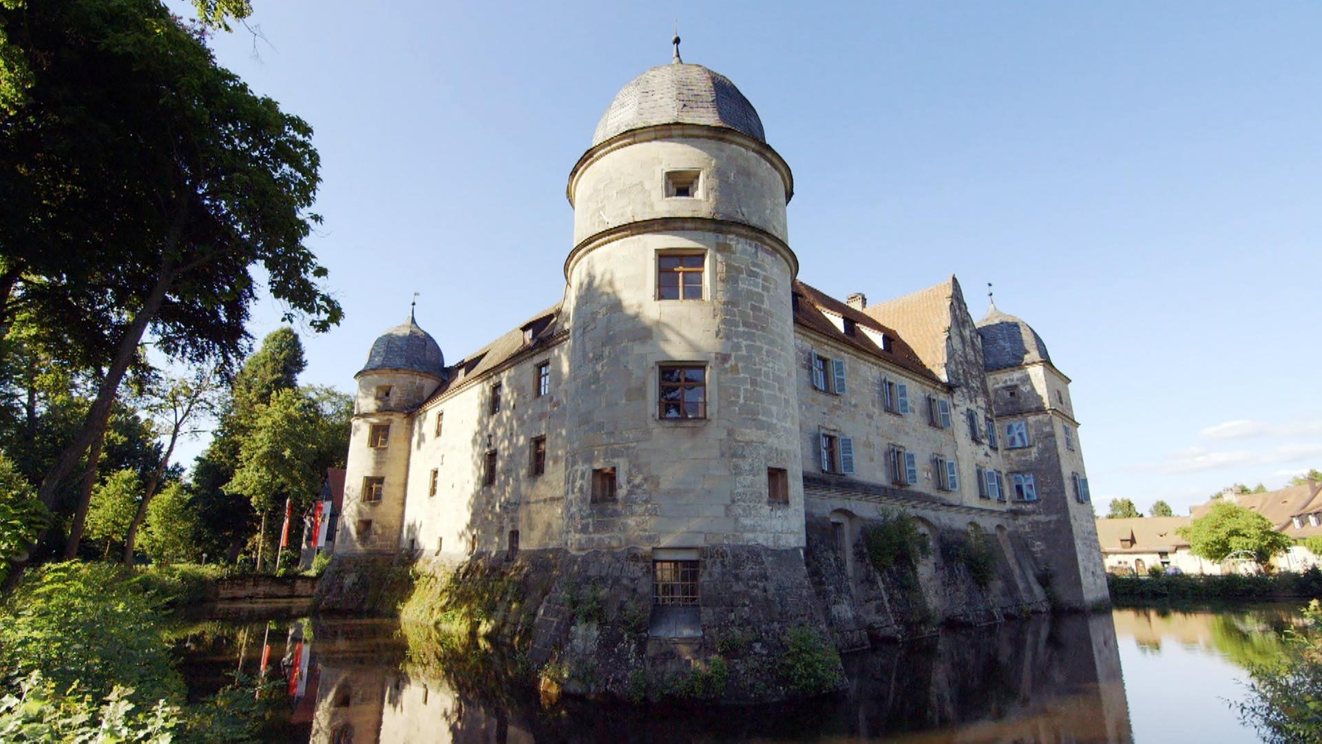 Ein Schloss umgeben von Wasser. Links davon sieht man Bäume, im Hintergrund sind Gebäude zu erkennen.