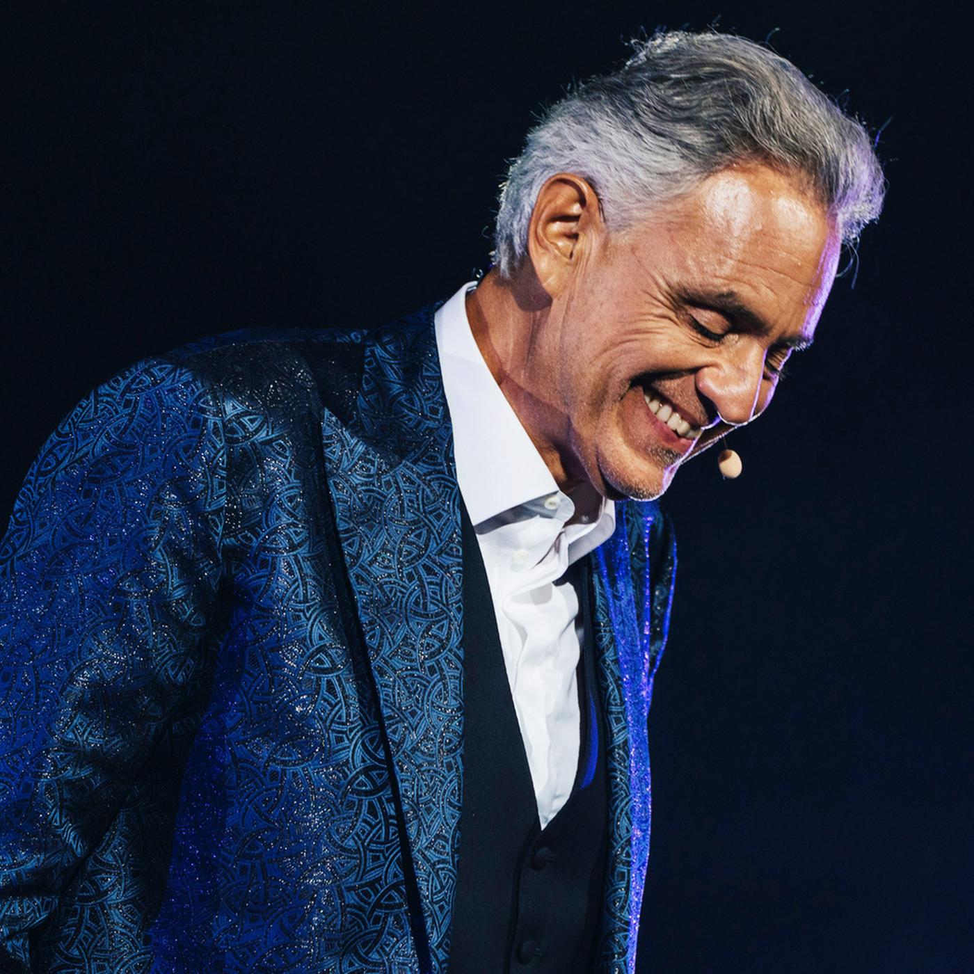 "Andrea Bocelli: Romanza": Das Bild zeigt eine einzelne Person auf einer Bühne vor einem dunklen, nahezu schwarzen Hintergrund. Die Szene ist durch gezielte Bühnenbeleuchtung in kühlen Blau- und Violetttönen ausgeleuchtet, die insbesondere die Konturen und die Kleidung hervorheben.