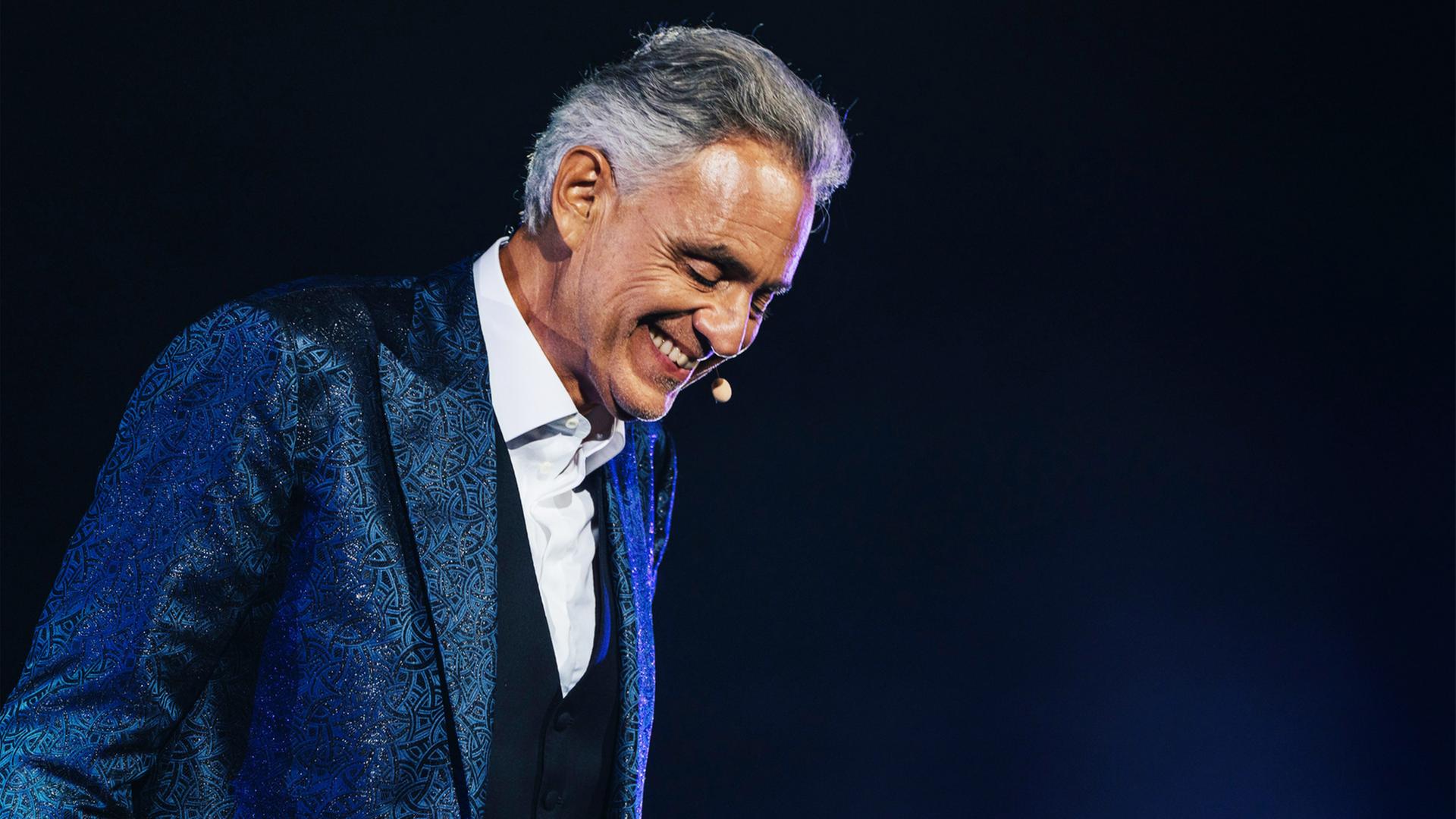 "Andrea Bocelli: Romanza": Das Bild zeigt eine einzelne Person auf einer Bühne vor einem dunklen, nahezu schwarzen Hintergrund. Die Szene ist durch gezielte Bühnenbeleuchtung in kühlen Blau- und Violetttönen ausgeleuchtet, die insbesondere die Konturen und die Kleidung hervorheben.
