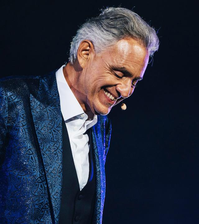 "Andrea Bocelli: Romanza": Das Bild zeigt eine einzelne Person auf einer Bühne vor einem dunklen, nahezu schwarzen Hintergrund. Die Szene ist durch gezielte Bühnenbeleuchtung in kühlen Blau- und Violetttönen ausgeleuchtet, die insbesondere die Konturen und die Kleidung hervorheben.