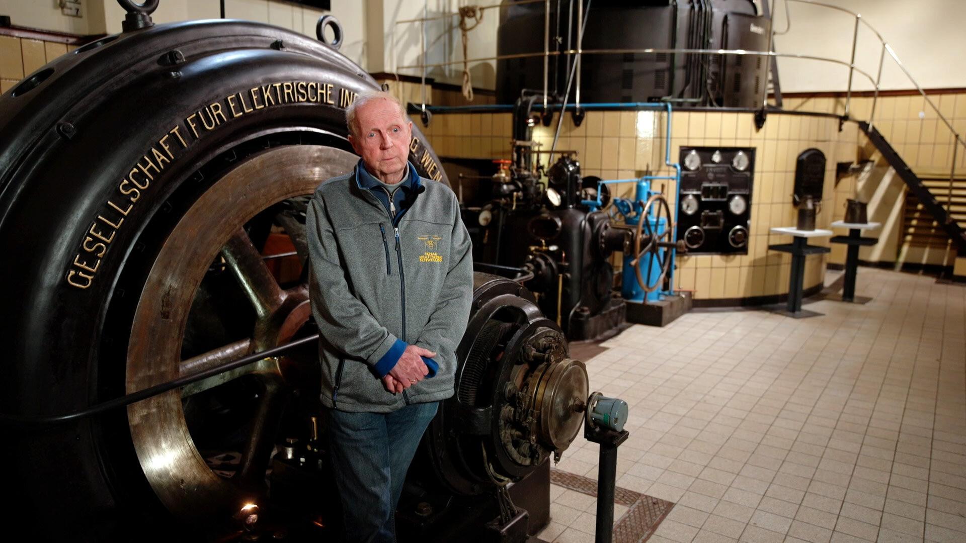 Eine historische Aufnahme zeigt Ernst Eibenberger in einer alten Kraftwerksanlage mit Maschinen im Vordergrund und Schaltanlagen im Hintergrund.