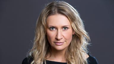 Das Bild zeigt eine Frau mit langen, welligen, blonden Haaren, die direkt in die Kamera schaut. Sie trägt ein einfaches schwarzes Oberteil und hat einen neutralen Gesichtsausdruck, der leicht selbstbewusst wirkt. Der Hintergrund ist einfarbig grau, was die Aufmerksamkeit auf ihr Gesicht lenkt. Es gibt keine weiteren Objekte oder Personen im Bild.   Die Beschreibung bezieht sich auf den Auftritt von Angelika Niedetzky beim Donauinselfest, wo sie mit Humor und einem scharfen Blick über alltägliche Themen spricht.