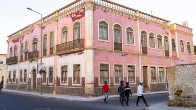 Angola: Vor einen heruntergekommenen Gebäude aus der Kolonialzeit läuft eine Gruppe schwarzer Männer über die Straße.