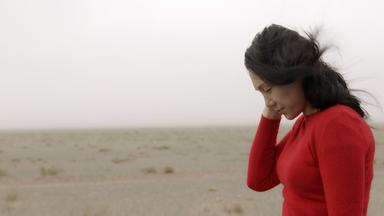 Ainur (seitlich fotografiert) steht in der mongolischen Wüste mit rotem Kleid und schwarzem Haar auf den Boden schauend.