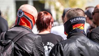 Foto zweier Männer in schwarzen Lederjacken von hinten. Einer ist kahlgeschoren und hat einen Schlauchschal mit Reichsflagge bis über die Nase hochgezogen. Der zweite ist kurzgeschoren und hat einen Schlauchschal mit Deutschlandfarben und dem Aufdruck Germany halb übers Gesicht gezogen.