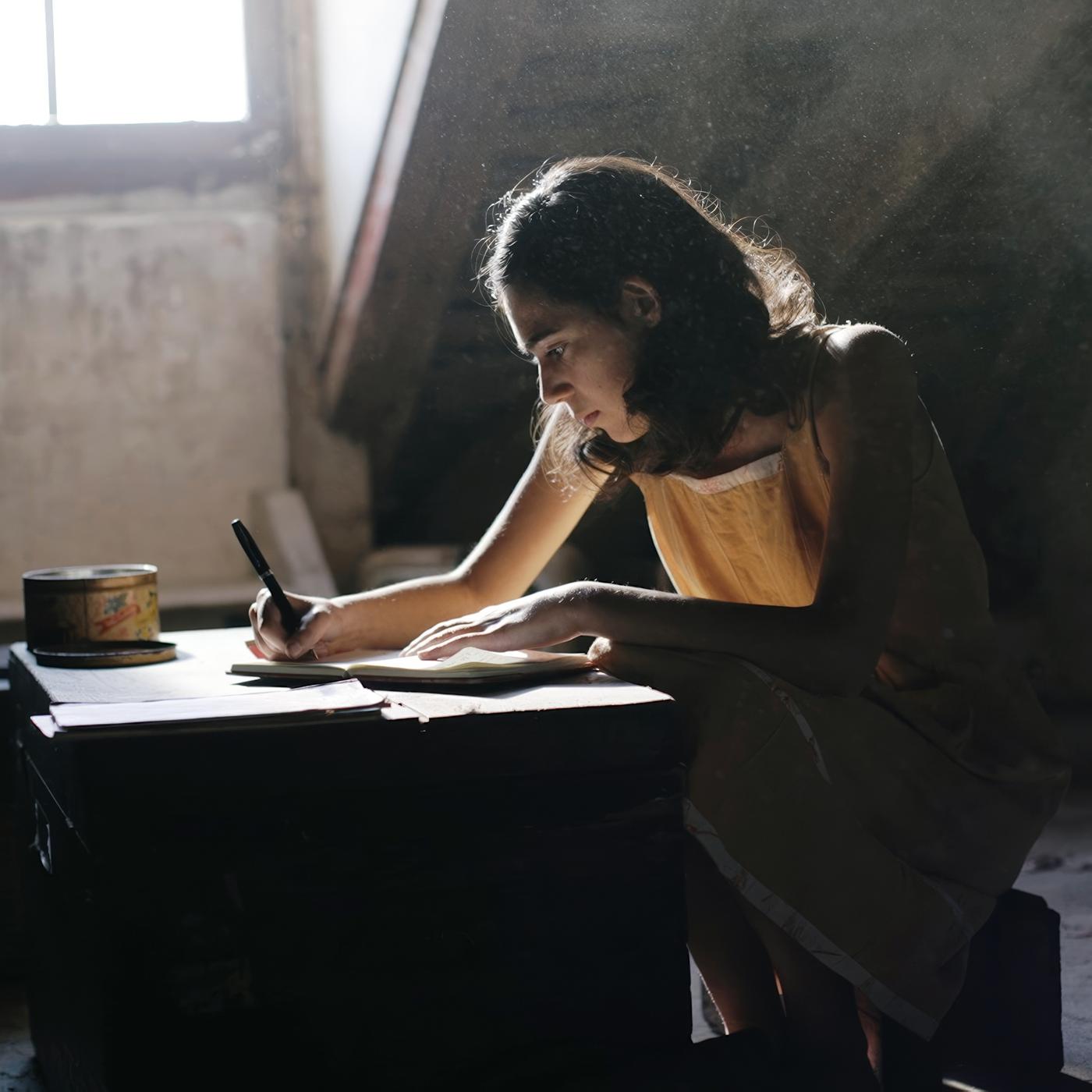 Das Bild in Schwarz-Weiß zeigt ein Mädchen an einem Schreibtisch, vor ihm liegt ein geöffnetes Buch und es hält einen Stift in der Hand. Das Mädchen trägt einen dunklen Pullover mit einer hellen Bluse darunter. Sie blickt neutral in die Kamera. Hinter ihr steht eine Topfpflanze.