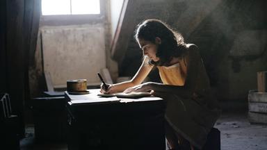 Das Bild in Schwarz-Weiß zeigt ein Mädchen an einem Schreibtisch, vor ihm liegt ein geöffnetes Buch und es hält einen Stift in der Hand. Das Mädchen trägt einen dunklen Pullover mit einer hellen Bluse darunter. Sie blickt neutral in die Kamera. Hinter ihr steht eine Topfpflanze.