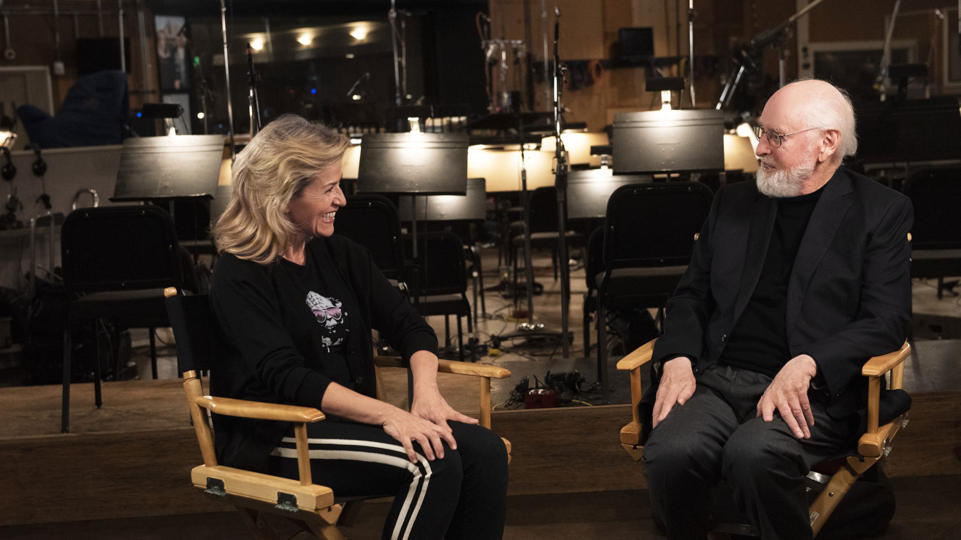 Anne-Sophie Mutter und John Williams sitzen im Bildvordergrund auf Regiestühlen. Sie tragen beide dunkle Kleidung und blicken sich an. Anne-Sophie Mutter (li) ist am lachen. Im Hintergrund sind Notenständer und Stühle in einem Tonstudio zu sehen.