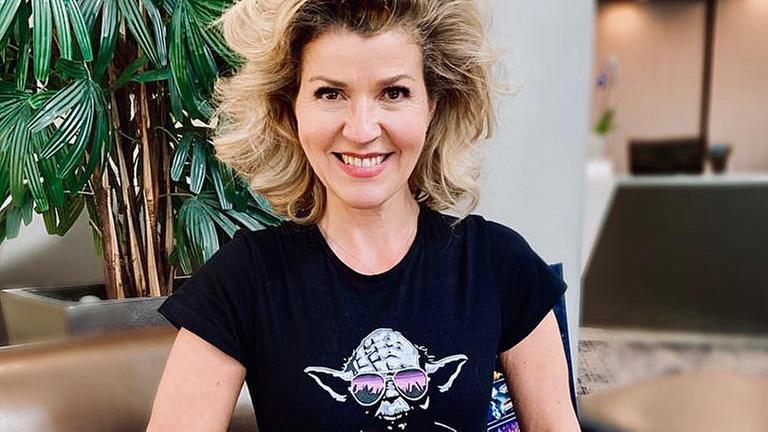 Anne-Sophie Mutter sitzt auffrecht auf einem braunen Ledersofa und lacht. Sie trägt ein schwarzes T-Shirt mit einem Meister Yoda Aufdruck. Dieser Trägt eine verspiegelte Sonnenbrille, um seinen Hals hängen Kopfhörer. Die Hände von Anne-Sophie Mutter umfassen den unteren Rand des T-Shirts und siehen es gerade.  