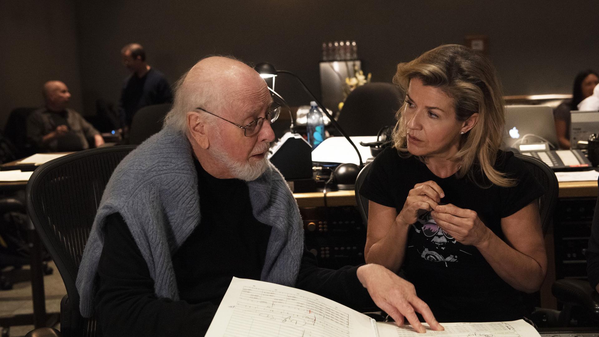 John Williams (li) und Anne-Sophie Mutter (re) sitzen vor einem Mischpult auf dem eine Partitur liegt. John Williams zeigt mit seiner linken Hand auf die Noten und tauscht sich mit Anne-Sophie Mutter aus. Im Hintergrund sind weiter Arbeitsplätze zu sehen.