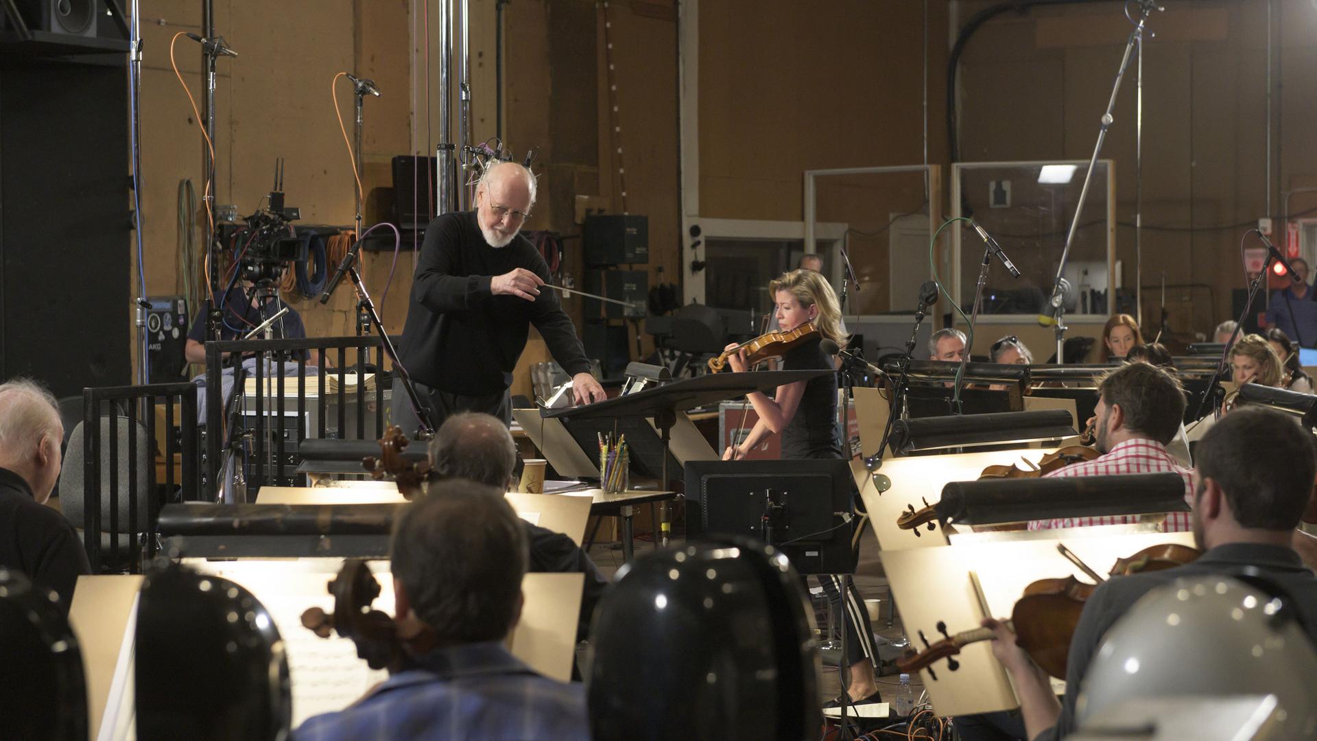 Anne-Sophie Mutter sitzt mit Orchestermusikern in einem Tonstudio. John Williams dirigiert diese Tonaufnahmen.