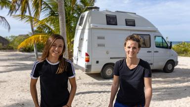 Anne (lange Haare, mit blauem Shirt und weißen Streifen) links, und Anna (mit blauem Shirt) rechts, vor Reisemobil am Strand