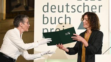Eine weißhaarige Frau überreicht einer anderen Frau mit Locken eine Kladde. Im Hintergrund steht "2020 deutscher Buchpreis"
