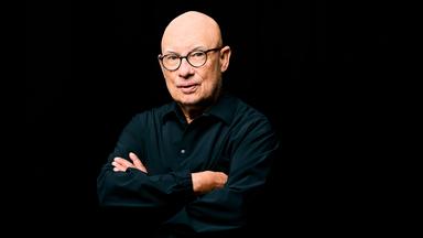 Ein Mann steht mit verschränkten Armen vor schwarzem Hintergrund.