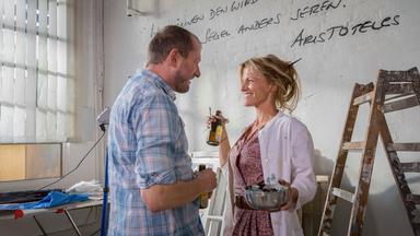 Maja (Janna Striebeck) steht ihrem Mann Florian (Martin Lindow) in seiner Malerwerkstatt gegenüber. Beide haben eine Flasche Bier in der Hand und lächeln sich an.