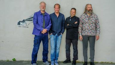 Vier Musiker stehen nebeneinander vor hellem Hintergrund; die Gruppe ist vollständig zu sehen und locker angeordnet.