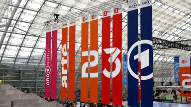 Vom Dach einer Glashalle hängen Banner mit den Logos von ARD, ZDF, 3sat und arte