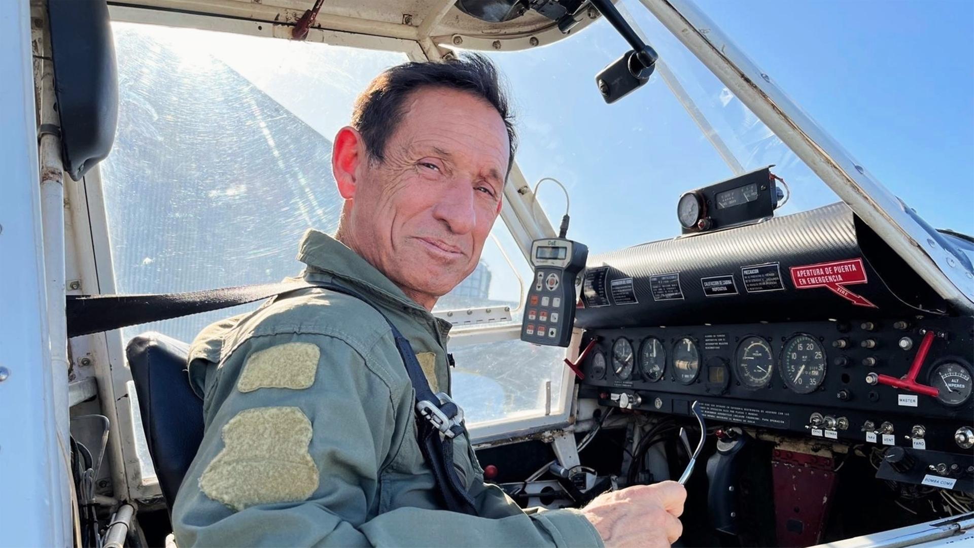 Ein älterer Mann sitzt in einem Flugzeugcockpit. Er trägt einen grünen Pilotenanzug mit Abzeichen. Vor ihm sind verschiedene Instrumente und Anzeigen. Im Hintergrund ist blauer Himmel zu sehen.