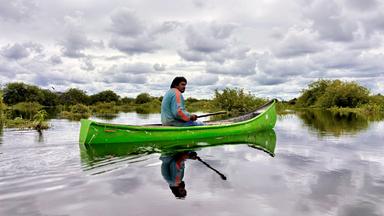 Ein Fischer sitzt in einem grünen Kanu auf ruhigem Wasser. Der Himmel ist von grauen Wolken bedeckt. Pflanzen und kleine Inseln sind im Hintergrund zu sehen.