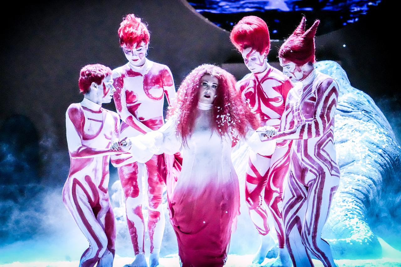 Das Bild zeigt eine Szene aus der Oper "Ariadne auf Naxos" von Richard Strauss, aufgeführt bei den Salzburger Festspielen 2026. Im Mittelpunkt steht eine Frau mit lockigem, rotem Haar, die ein langes, weißes Gewand trägt. Sie scheint in einer verletzlichen Pose zu stehen und wird von vier Personen umgeben, die in engen, bunten Körperanzügen gekleidet sind. Diese Anzüge sind weiß mit roten und pinken Mustern, und alle Figuren haben auffällige, rote Frisuren, die in verschiedenen Formen gestylt sind. Die Kulisse wirkt mystisch mit gedämpfter Beleuchtung und einem blauen, sanften Hintergrund, der auf Wasser anspielt. Der gesamte Eindruck des Bildes vermittelt eine Mischung aus Tragik und Komik, passend zur thematischen Tiefe der Aufführung.