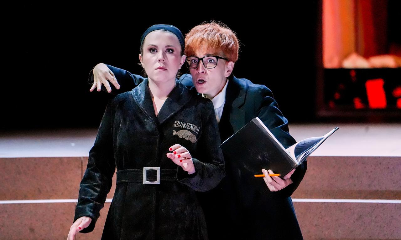 Das Bild zeigt eine Szene aus der Oper "Ariadne auf Naxos" von Richard Strauss, die während der Salzburger Festspiele 2026 aufgeführt wurde. Im Vordergrund stehen zwei Figuren. Die erste Figur ist eine Frau mit einem ernsthaften Gesichtsausdruck, die in einem schwarzen, samtartigen Outfit gekleidet ist, das einen breiten Gürtel hat. Sie trägt ein schwarzes Stirnband und hat ihre Augenbrauen zusammengezogen, was ihre Verwunderung oder Besorgnis ausdrücken könnte.  Die zweite Figur ist eine männlich wirkende Person mit kurz geschnittenem, orangefarbenem Haar und einer auffälligen Brille. Der Ausdruck dieser Figur ist energetisch und überrascht; sie hält ein aufgeschlagenes Buch in einer Hand und einen Stift in der anderen. Die linke Hand der ersten Figur ist auf die Schulter der zweiten Figur gelegt, was eine Verbindung oder Interaktion zwischen beiden darstellt.  Der Hintergrund des Bildes ist dunkel und es sind erleuchtete Elemente sichtbar, die auf eine theatrale Kulisse hinweisen, welche möglicherweise einen dramatischen oder komischen Anteil an der Aufführung widerspiegelt.