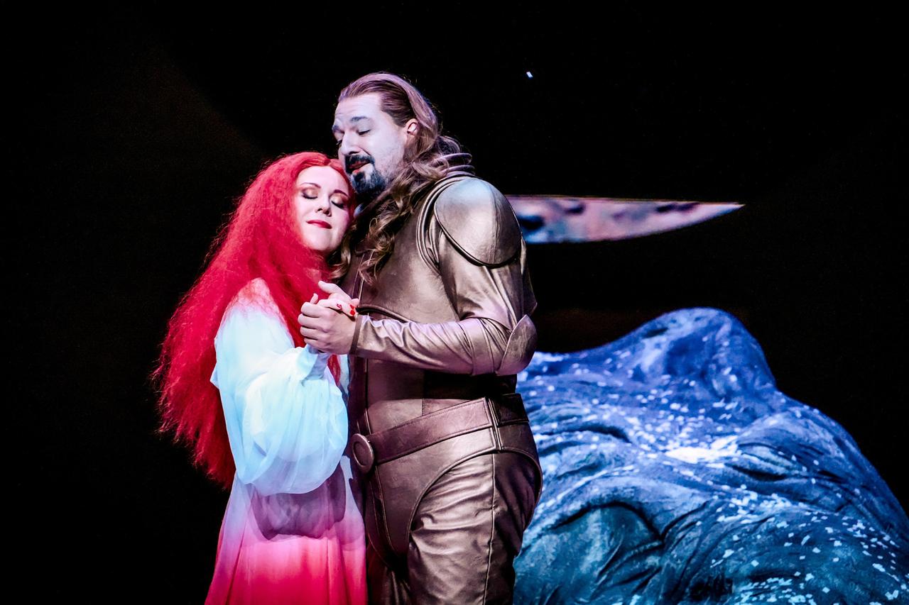 Das Bild zeigt eine Szene aus der Oper "Ariadne auf Naxos" von Richard Strauss, die während der Salzburger Festspiele 2026 aufgeführt wurde.   In der Mitte stehen zwei Hauptfiguren. Die erste Figur ist eine Frau mit auffallend roten, langen Haaren, die ein fließendes, leicht transparentes Kleid trägt, das von Weiß zu Rosa verläuft. Sie steht eng umschlungen mit der zweiten Figur, einem Mann, der in einem glänzenden, goldfarbenen Kostüm mit Rüstungsteilen gekleidet ist. Die beiden Figuren schauen sich an und scheinen in einem emotionalen Moment des Zusammenhalts zu sein.  Im Hintergrund ist eine dunkle, unifarbene Fläche zu sehen, die die Bühne darstellt. Eine große, blaue, steinartige Masse ist teilweise sichtbar, was wahrscheinlich eine dekorative Kulisse der Insel Naxos darstellt. Der Szenenaufbau vermittelt eine dramatische und gefühlvolle Atmosphäre, die die Inhalte der Oper widerspiegelt, in der Konflikte und Transformationsprozesse thematisiert werden.