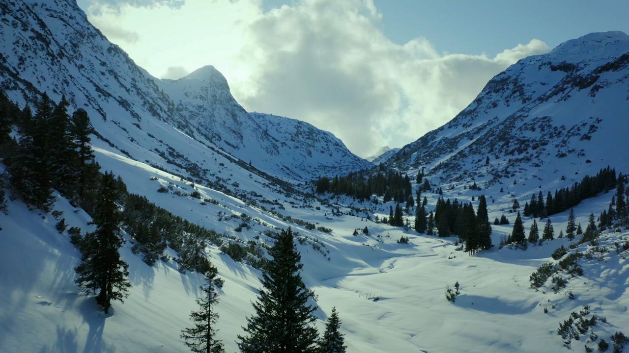 Das Bild zeigt eine winterliche Berglandschaft im Arlberg. Schneebedeckte Hänge und spitze Felsen bestimmen die Szenerie. Im Vordergrund stehen Kiefern, während der Himmel teilweise bewölkt ist.