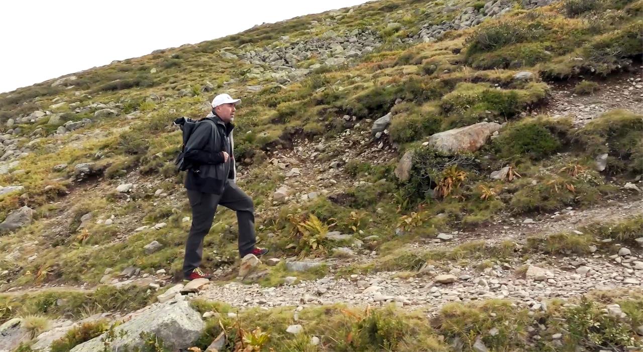 Das Bild zeigt einen Mann, der auf einem schmalen Wanderweg in einer alpinen Landschaft geht. Er trägt eine leichte Jacke, eine Mütze und hat einen Rucksack auf dem Rücken. Der Weg ist steinig und von Gras und kleinen Pflanzen umgeben. Im Hintergrund sind Hügel und eine wolkenverhangene Straße sichtbar, die die natürliche Umgebung prägen. Der Mann scheint aktiv und konzentriert auf seinem Weg zu sein. Die Landschaft ist typisch für alpine Regionen, mit sanften Erhebungen und sporadischer Vegetation.