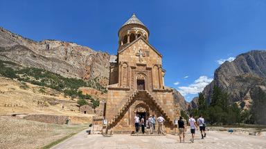 Das Bild zeigt die Klosterkirche Noravank in Armenien, umgeben von einer felsigen Landschaft. Mehrere Personen betreten die Kirche über eine Treppe.