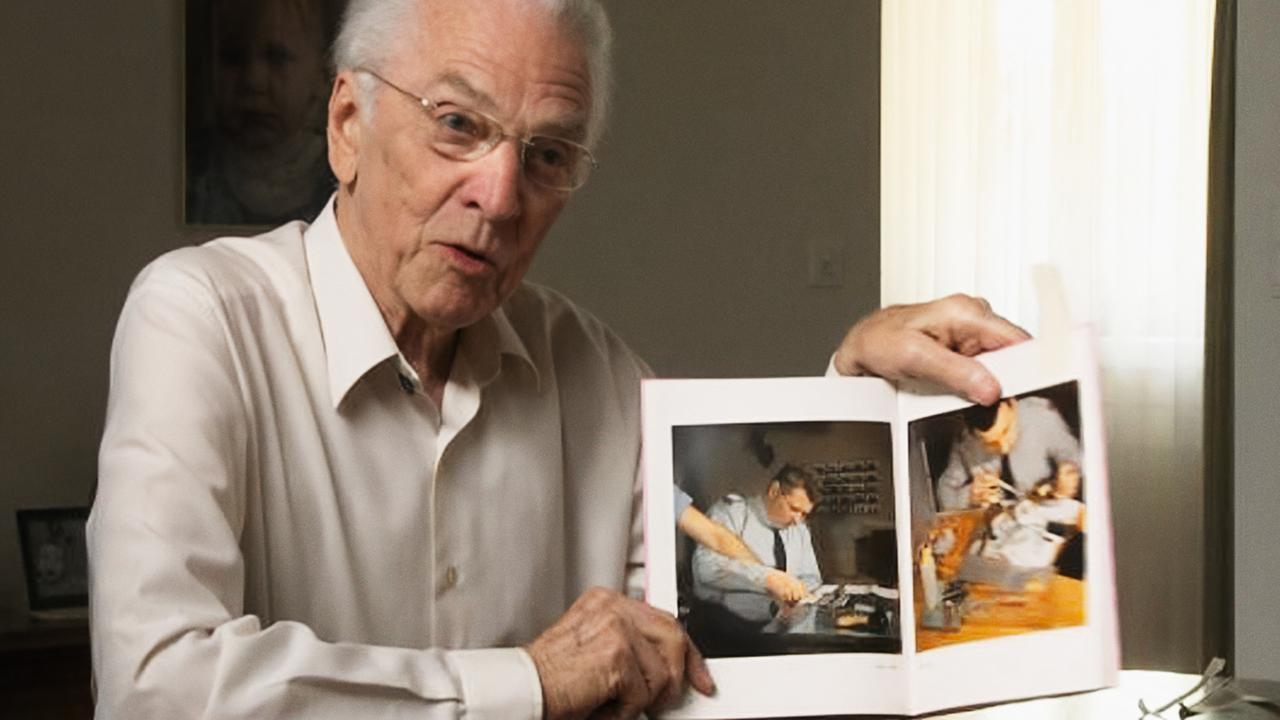 Ein älterer Mann mit weißem Haar und Brille sitzt an einem Tisch und zeigt in einem aufgeschlagenen Buch Fotografien von Polizeiarbeit.