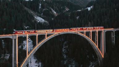 Ein roter Zug fährt in der Dämmerung über eine beleuchtete Brücke, umgeben von einer winterlichen Berglandschaft mit schneebedeckten Gipfeln.