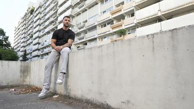 Ein junger Mann sitzt auf einer Betonwand vor einem Hochhaus