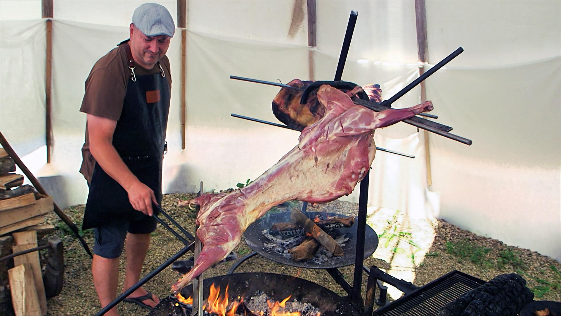 Das Bild zeigt einen Mann, der als Jäger-Asador bezeichnet wird. Er steht vor einem offenen Feuer und verwendet lange Grillzangen, um ein großes, am Spieß hängendes Stück Fleisch zu wenden. Das Fleisch ist gut sichtbar und wird über der Flamme gegrillt. Im Hintergrund sieht man eine einfache Zeltstruktur, die aus weißen Plane besteht, und am Boden liegt ein Kies- und Schotterboden. Vom Feuer geht eine warme, rauchige Atmosphäre aus, die die Szene prägt. Es sind auch einige Holzscheite sichtbar, die um das Feuer herum liegen. Die Umgebung wirkt rustikal und naturverbunden, passend zur Thematik des traditionellen Grillens.