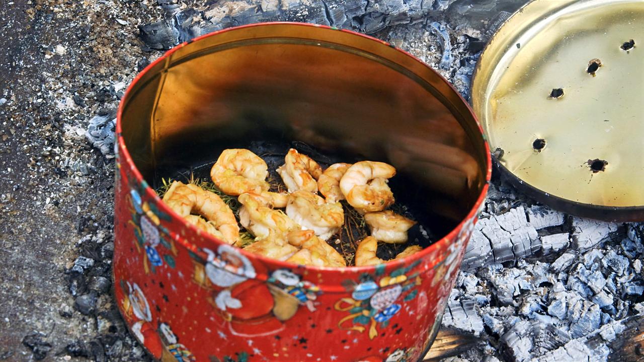 Das Bild zeigt eine rote Keksdose, die geöffnet ist und in der sich mehrere gegrillte Garnelen befinden. Die Garnelen sind goldbraun gebraten und liegen auf etwas grünem Gras oder Kräutern. Über der Dose ist der Deckel, der ebenfalls rot ist und mit einem goldenen Rand sowie kleinen Löchern versehen ist. Der Hintergrund besteht aus grauen, verkohlten Holzstücken und Asche, die von einem offenen Feuer stammen. Die gesamte Szene vermittelt eine rustikale Atmosphäre und zeigt eine traditionelle Grillmethode.