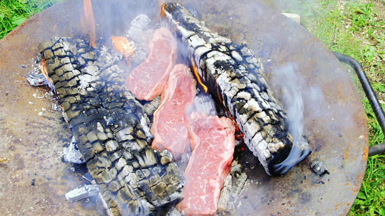 Das Bild zeigt eine Grillstelle mit mehreren Stücken Fleisch, die direkt über glühenden Kohlen gegrillt werden. Die Kohlen sind teilweise verbrannt und erzeugen Rauch, der in die Luft aufsteigt. Das Grillgerät ist eine flache, runde, metallene Fläche, auf der das Fleisch liegt. Im Hintergrund sind Rasenflächen und einige unscharfe, grüne Elemente zu erkennen. Das Gesamtbild vermittelt eine rustikale Atmosphäre, die auf traditionelle Grillmethoden hindeutet.