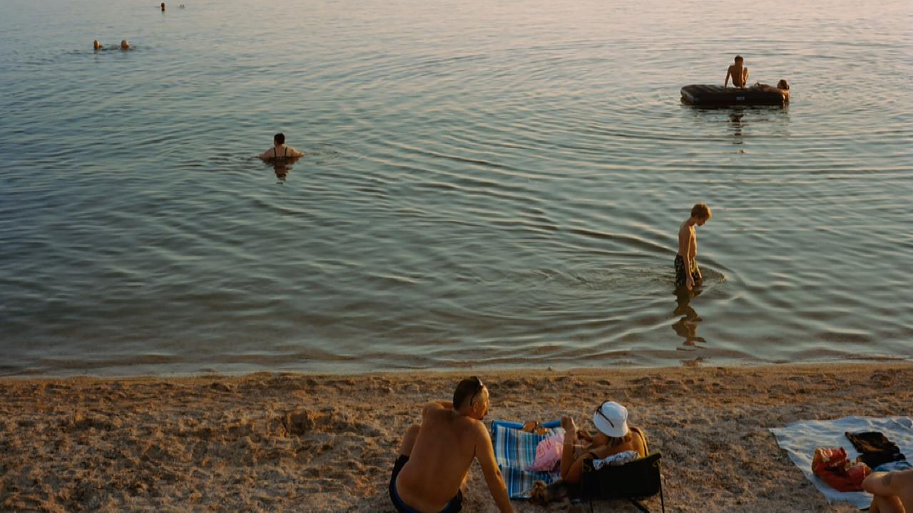Menschen liegen am Strand in der Sonne, während andere im Meer schwimmen, oder auf Schlachbooten unterwegs sind.