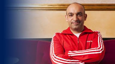 Der Kabarettist Django Asül sitzt in einem Trainingsanzug vor einer Wand.