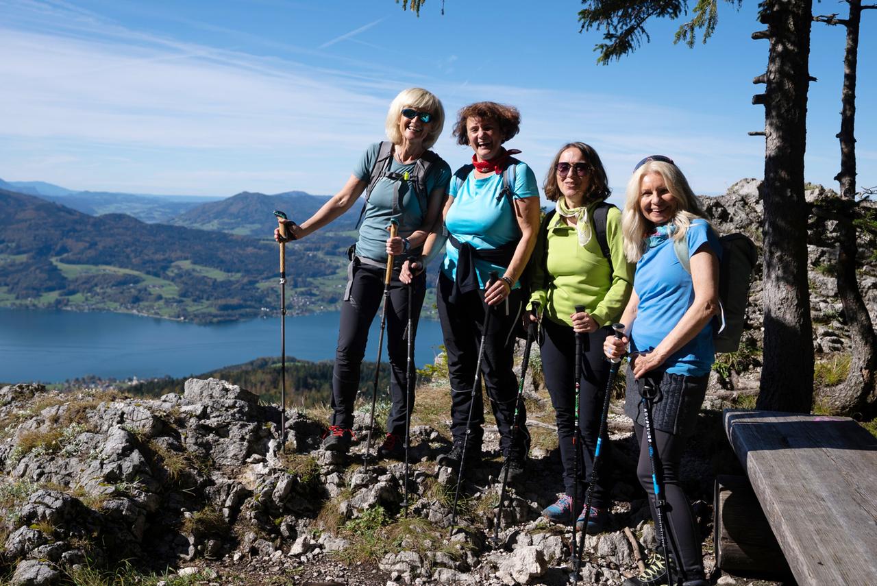 Das Bild zeigt vier Frauen, die auf einem felsigen Aussichtspunkt stehen. Die Frauen sind in sportlicher Kleidung und halten Wanderstöcke in der Hand. Im Hintergrund ist eine weitläufige, malerische Landschaft mit grünen Hügeln und einem großen, blauen See zu sehen, der wahrscheinlich der Attersee ist. Der Himmel ist klar und blau, und die Umgebung wirkt sonnig. Einige Bäume und Felsen umrahmen die Szene. Die Frauen lächeln und scheinen eine positive Stimmung auszustrahlen.