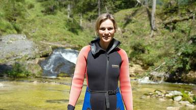 Das Bild zeigt eine Frau, die in einem Gewässer steht, umgeben von einer grünen, bewaldeten Landschaft. Sie trägt einen schwarzen Wetsuit mit roten und blauen Akzenten sowie Handschuhe. Hinter ihr sind klare Wasserläufe und kleine Kaskaden zu sehen, die sanft über Steine fließen. Der Hintergrund ist von Bäumen und Hügeln geprägt, die eine natürliche, idyllische Umgebung schaffen. Das Licht wirkt hell und freundlich, was auf einen sonnigen Tag hindeutet.