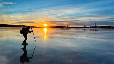 Eine Person mit Rucksack und auf Schlittschuhen läuft auf einem zugefrorenen See. Im Hintergrund geht die Sonne unter.