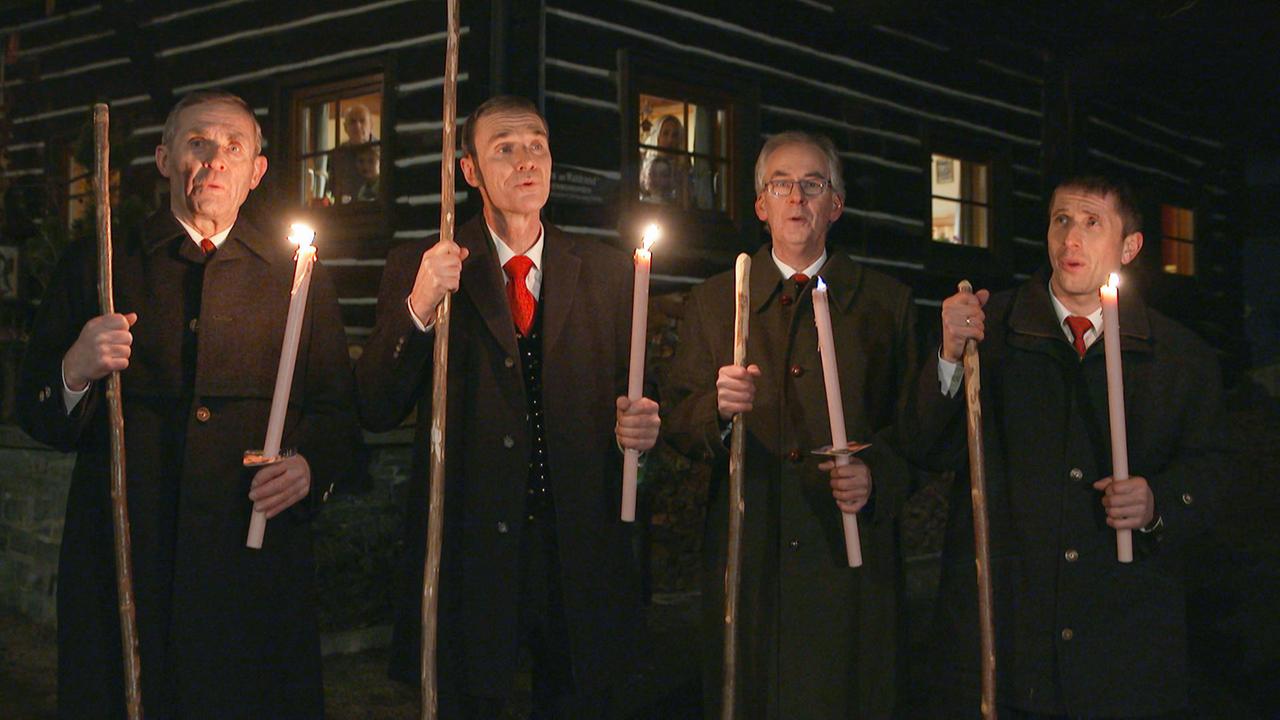 Vier Sänger mit Stäben und langen Kerzen vor einer Hütte