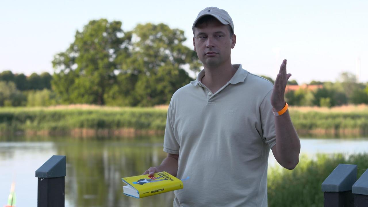 Mann mit Basecap vor Gewässer hält gelbes Buch im Hand und gestikuliert.