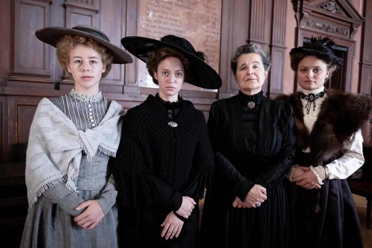Das Bild zeigt vier Frauen, die in historischen Kleidern aus der Zeit um 1900 gekleidet sind. Sie stehen nebeneinander in einem Raum mit holzverkleideten Wänden und großen Fenstern. Die Frauen haben unterschiedliche Outfits: 

1. Die linke Frau trägt ein graues Kleid mit einer weißen, gestrickten Stola. 
2. Die zweite von links trägt ein schwarzes, aufwändiges Kleid und hat einen großen schwarzen Hut mit einer Blume.
3. Die dritte Frau zeigt ein schlichtes schwarzes Outfit und hat graue Haare, die zu einem strengen Dutt frisiert sind.
4. Die rechte Frau trägt ein weißes Oberteil mit einer dunklen Rockgarnitur und hat einen Pelzkragen.

Alle Frauen haben ernste Gesichtsausdrücke und stehen mit verschränkten Armen. Der Raum, in dem sie sich befinden, hat eine historische Atmosphäre mit rustikalen Details. Es gibt im Hintergrund eine Holztafel mit Inschriften.