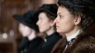 Das Bild zeigt eine Szene aus einem historischen Films. Im Vordergrund sieht man eine Frau in einem eleganten, historischen Kostüm, die zur Seite schaut. Sie trägt einen dunklen Hut und hat ihre Haare hochgesteckt. Ihr Outfit ist verziert mit einer auffälligen Halskette. Hinter ihr sind zwei weitere Frauen sichtbar, die ebenfalls ähnliche historische Kleidung tragen, jedoch nur teilweise erkennbar sind, da ihre Gesichter zum Teil im Schatten liegen.

Die Umgebung scheint ein geräumiger Innenraum zu sein, möglicherweise ein Gerichtssaal oder ein ähnlicher Ort, der mit dunklem Holz verkleidet ist. Die Stimmung des Bildes kann als ernst und nachdenklich beschrieben werden, passend zum Kontext des Films, der sich mit Themen wie Frauenhandel und gesellschaftlichen Missständen zu Beginn des 20. Jahrhunderts beschäftigt.