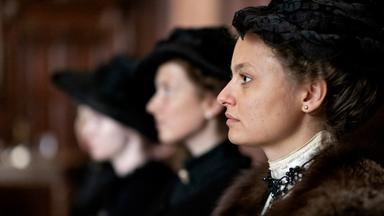 Das Bild zeigt eine Szene aus einem historischen Films. Im Vordergrund sieht man eine Frau in einem eleganten, historischen Kostüm, die zur Seite schaut. Sie trägt einen dunklen Hut und hat ihre Haare hochgesteckt. Ihr Outfit ist verziert mit einer auffälligen Halskette. Hinter ihr sind zwei weitere Frauen sichtbar, die ebenfalls ähnliche historische Kleidung tragen, jedoch nur teilweise erkennbar sind, da ihre Gesichter zum Teil im Schatten liegen.  Die Umgebung scheint ein geräumiger Innenraum zu sein, möglicherweise ein Gerichtssaal oder ein ähnlicher Ort, der mit dunklem Holz verkleidet ist. Die Stimmung des Bildes kann als ernst und nachdenklich beschrieben werden, passend zum Kontext des Films, der sich mit Themen wie Frauenhandel und gesellschaftlichen Missständen zu Beginn des 20. Jahrhunderts beschäftigt.