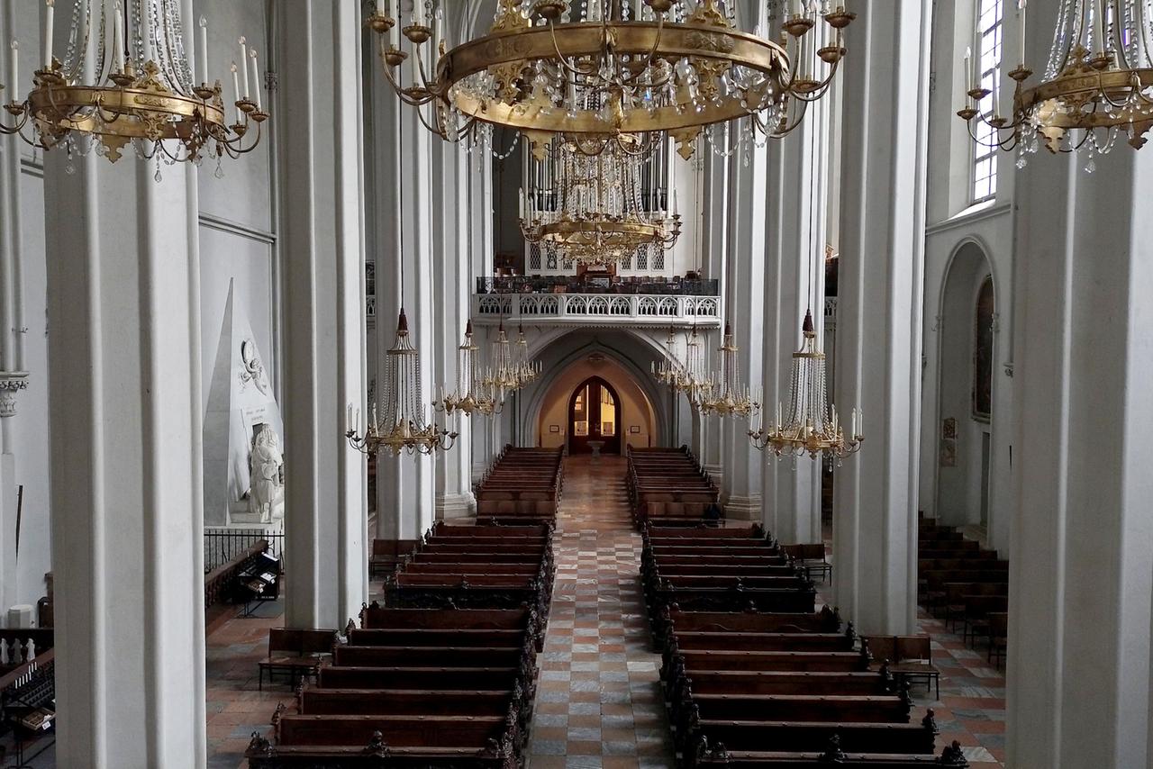Innenansicht der Augustinerkirche in Wien mit Blick auf den Mittelgang, flankiert von Bänken und Kronleuchtern, führend zu einem beleuchteten Bogen am Ende.